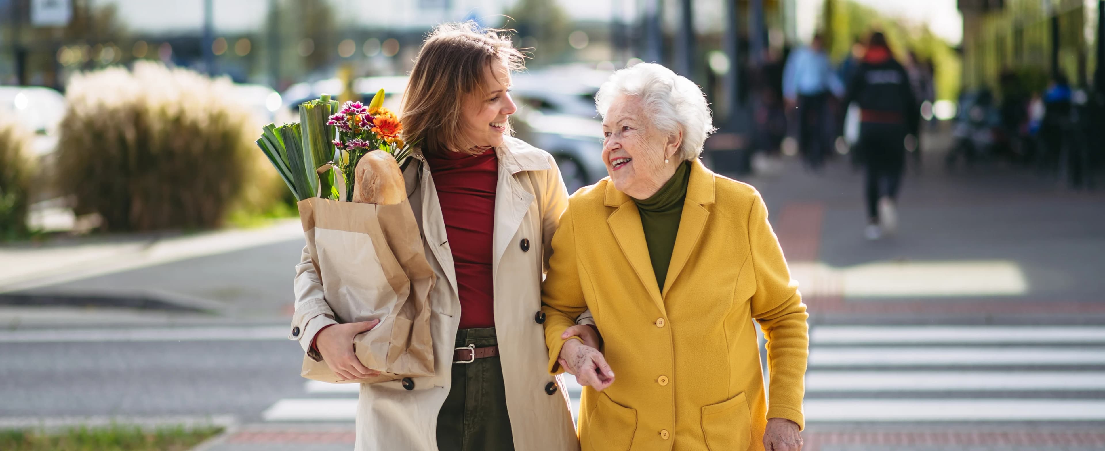 Fröhliche ältere Dame und ihre Pflegekraft gehen mit Einkäufen die Straße entlang