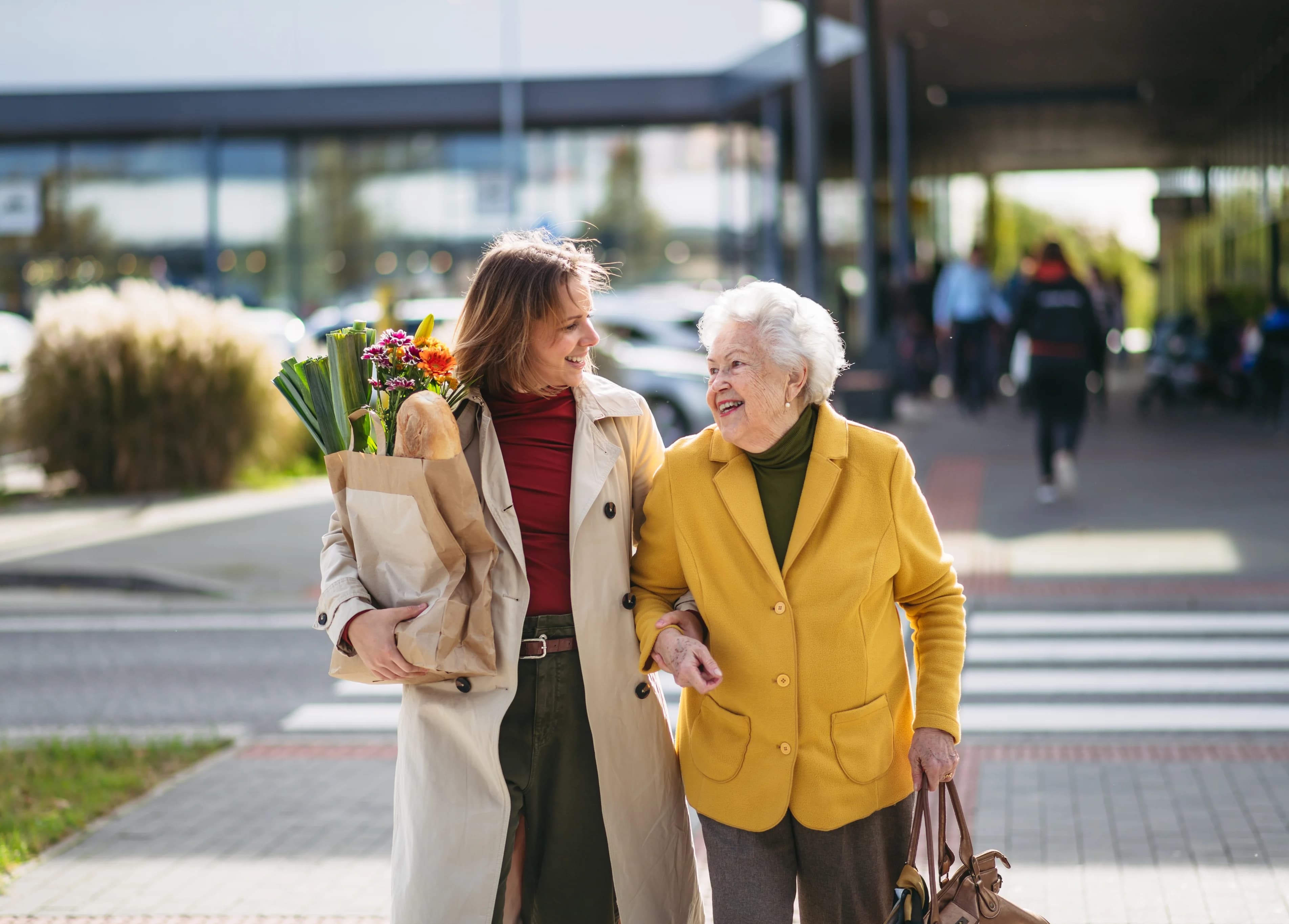 Fröhliche ältere Dame und ihre Pflegekraft gehen mit Einkäufen die Straße entlang
